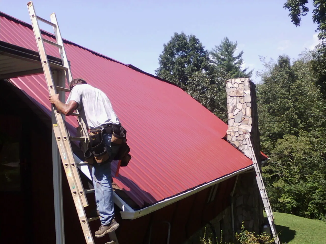 Roofer on a ladder beside a red metal roof and stone chimney — Soffit & Fascia Repair by Roofing Brothers in Amanda Park